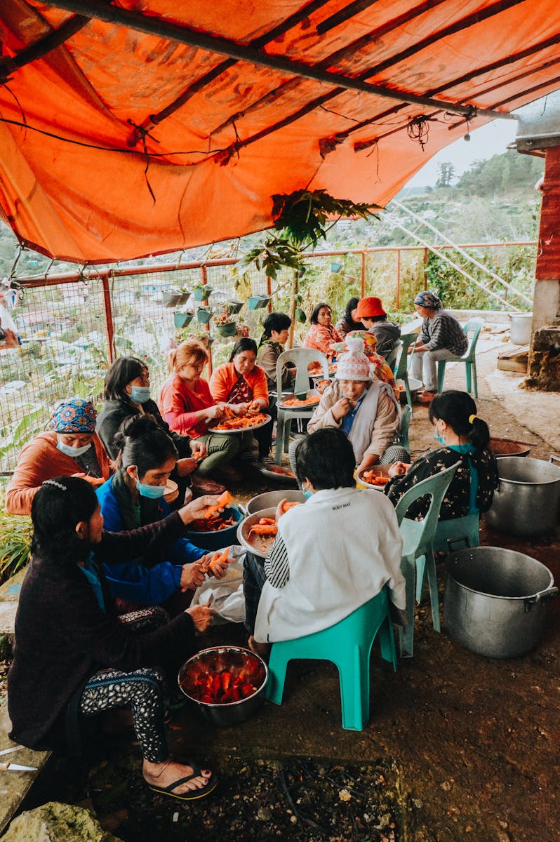 A group of people sitting together outdoors, peeling and preparing vegetables.