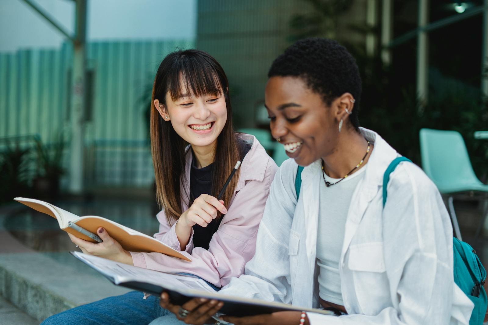 Young positive multiethnic female students with exercise books talking while doing homework on street stairs