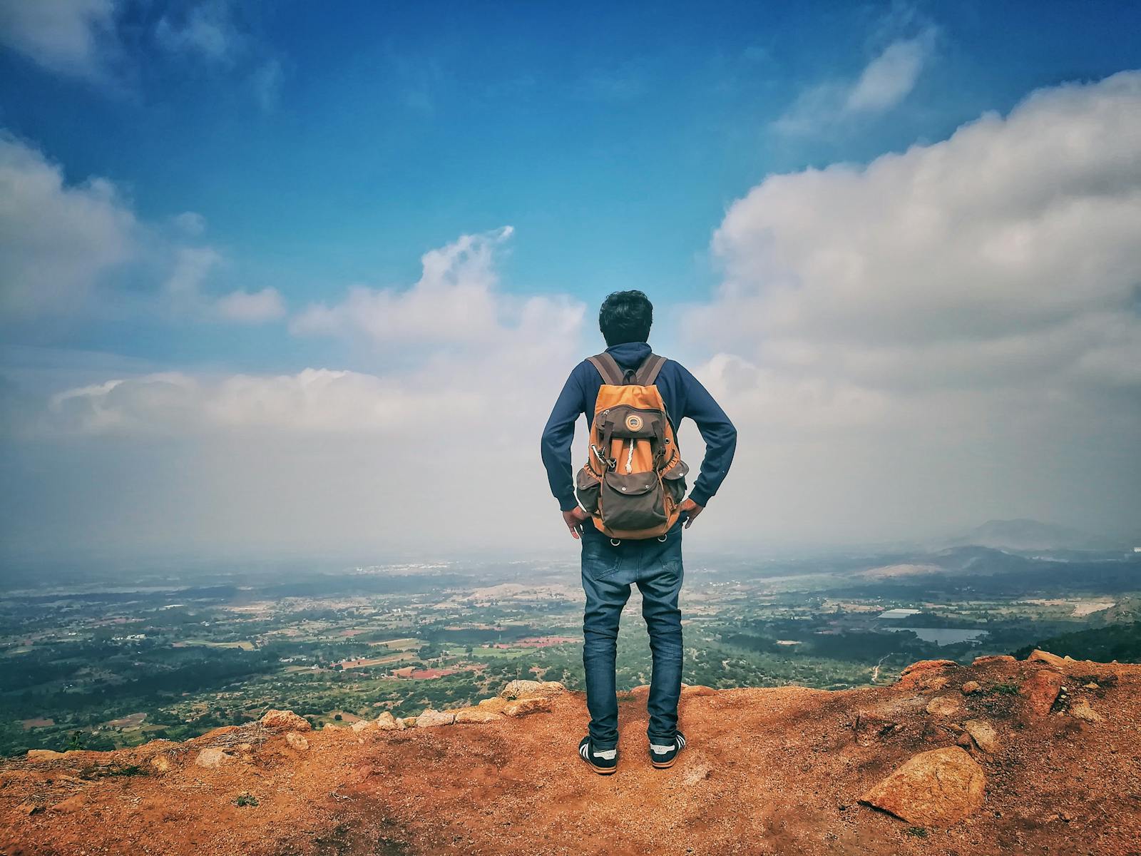 A man stands on a mountain edge appreciating the expansive scenic view below under a bright sky.